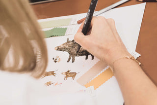 Eine Person, die auf weißem Papier Wildschweine mit verschiedenen farbigen Markern auf Stoffstreifen zeichnet, wobei sie sich auf Details konzentriert.