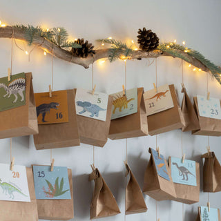 DIY-Adventskalender-Set Dinosaurier mit nummerierten Dinosaurier-Karten auf braunen Papiertüten, an Ast mit Lichtern hängen.