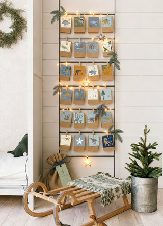 Ein weihnachtliches Kinderzimmer im skandinavischen Stil zeigt einen DIY Adventskalender mit Dinosaurierkarten und Kraftpapierbeutel. Diese sind dekorativ an einer Metallleiter befestigt und mit Zweigen und Lichterkette hübsch dekoriert.