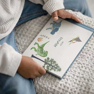Kind mit einem „Dino-Freundebuch“ mit bunten Dinosaurier-Illustrationen auf dem Einband, bekleidet mit einem gemütlichen Pullover und Jeans.
