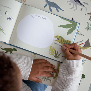 Ein Kind zeichnet in einem Dino-Freundebuch, umgeben von bunten Dinosaurier-Illustrationen und Namen. Fokus auf Triceratops Bild.