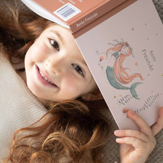 Ein fröhliches Kind liegt mit dem "Freundebuch Beste Freunde" in der Hand, das eine Meerjungfrau und ein Einhorn auf dem Cover zeigt.