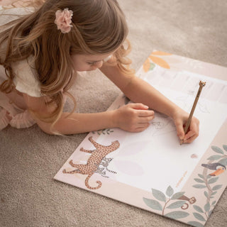 Ein Kind malt auf einer Papierblockunterlage verschiedene Tiere. Auf der Kinderschreibtischunterlage sind viele verschiedene, von Hand gezeichnete Jungle Tiere dargestellt. Die Farben sind zartrosa, apricot, grün und beige.