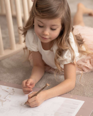 Ein Mädchen malt Figuren auf eine Papierblockunterlage. Die Kinderschreibtischunterlage zeigt  ein magisches weißes Einhorn welches fröhlich vor dem zarten,  transparenten Märchenschloss springt.