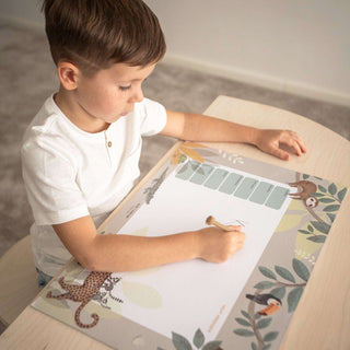 Ein Kind sitzt am Kinderschreibtisch und malt Buchstaben auf eine Papierblockschreibtischunterlage. Auf der Unterlage sind Jungle Tiere zu sehen. Die Hintergrundfarbe ist taupe mit verschieden Grüntönen.