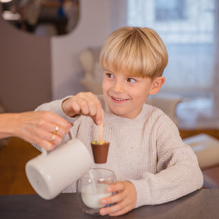Ein Kind bekommt Milch in eine Tasse gegossen. Es hält lächelnd eine feste Trinkschokolade am Holzstil über die Tasse.
