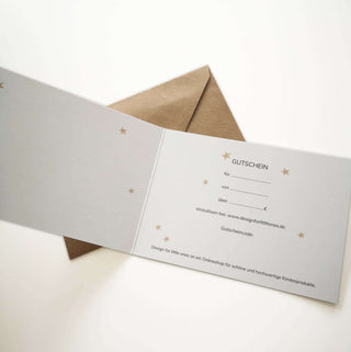 Ein weißer Geschenkgutschein mit Sternenmustern, geöffnet, um Felder zum Ausfüllen zu zeigen, begleitet von einem braunen Umschlag.