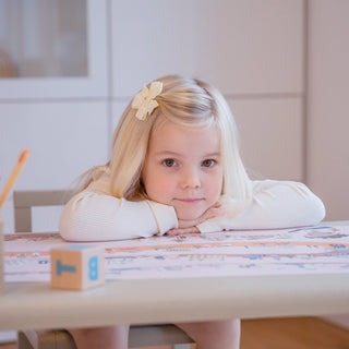 Lächelndes Mädchen an Kinderschreibtisch mit rosa Zeitreise Unterlage: Glückliches Kind schaut frontal in die Kamera, sitzend an einem Tisch mit der pädagogischen rosa Schreibtischunterlage "Zeitreise der Menschheit". Ideal für kleine Entdeckerinnen.