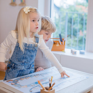 Kind zeigt auf Schreibtischunterlage Zeitreise: Blondes Mädchen mit blauer Latzhose entdeckt spielerisch die Illustrationen der Menschheitsgeschichte auf der blauen Lernunterlage für den Kinderschreibtisch.