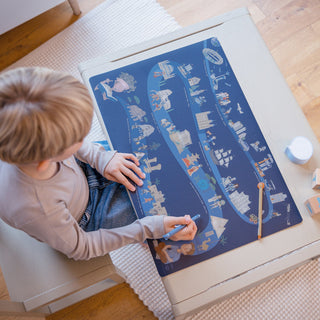 Kind sitzt von oben gesehen an einem Tisch, malt mit einem Stift auf einem Blatt Papier, die navy-blaue Schreibtischunterlage "Zeitreise der Menschheit" liegt darunter.