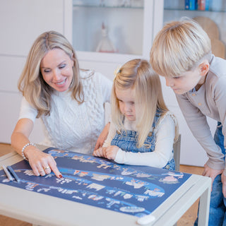 Frau und zwei Kinder, ein Mädchen und ein Junge, beugen sich über die navy-blaue Schreibtischunterlage "Zeitreise der Menschheit" und entdecken gemeinsam die Illustrationen.