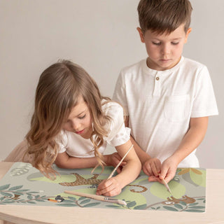 Zwei Kinder stehen hinter einem Kinderschreibtisch und malen mit Pinseln auf einer Schreibtischunterlage mit Dschungelmotiv.