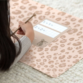 Das Kind schreibt auf einer abwaschbaren, leopardengemusterten Schreibunterlage in zartem Beige mit braunen Flecken, die auf einer leicht strukturierten Oberfläche liegt.