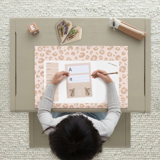 Kind am Tisch mit Papierblock-Unterlage im Leopardenmuster, Wochenplaner, Schreibutensilien und Buchstaben-Übungsblatt.