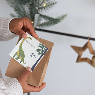 Adventskalender-Set mit Dinosaurier-Karte und brauner Papiertüte, Hände halten Karte mit „Brachiosaurus“ und Nummer 14.
