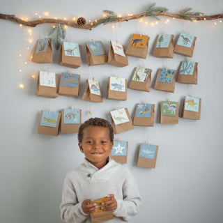 Ein Kind steht vor einem Dinosaurier-Adventskalender-Set. Bunte, nummerierte Beutel mit Dinosauriermotiven hängen an einem dekorierten Ast.