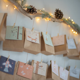 DIY Adventskalender Set mit braunen Papiertüten und kindgerechten Motiven wie einem Fuchs, aufgehängt mit Wäscheklammern. Der weihnachtliche Adventskalender ist an einem Ast mit Tannenzapfen und Lichterkette dekoriert und schafft eine gemütliche Festtagsstimmung.