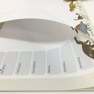 Ein Wochenplaner mit Dinosaurier-Illustrationen, die mit den Wochentagen beschriftet sind und auf einer Tischplatte liegen.
