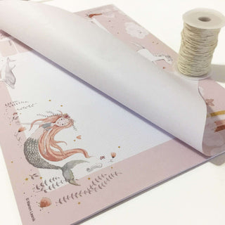Papierblock Unterlage im Meerjungfrau-Design mit pastellrosa Wochenplaner, illustrierten Meerjungfrauen, Meereselementen, neben Garnrolle.