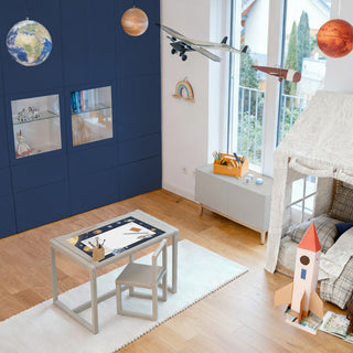 Kinderzimmer im Weltallstil mit Planeten und Rakete. In der Mitte steht ein Kinderschreibtisch mit einer blauen Papierblockunterlage mit Weltalldesign.