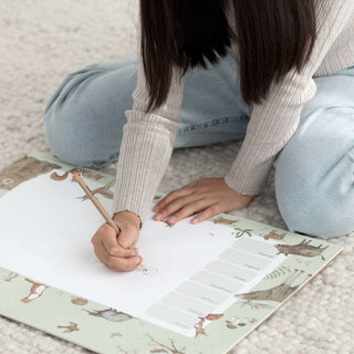 Mädchen mit „Waldtiere grün“-Wochenplaner mit Waldtiermotiven, schreibt mit einem Bleistift, während sie auf einem gemütlichen Teppich sitzt.