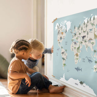 Zwei Kinder sitzen im Kinderzimmer auf dem Boden und schauen ganz neugierig und interessiert ein Poster mit einer Dino-Weltkarte an.