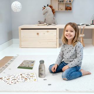Ein Mädchen sitzt glücklich im Kinderzimmer auf dem Boden. Sie hat eine Kindertrinkflasche mit Tier-Stickern beklebt und ist nun fertig.