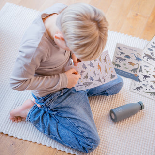 Ein Kind sitzt auf dem Boden und ist dabei ein Dino-Sticker vom Bogen abzulösen. Das gesamte Sticker-Set für Kinder liegt daneben auf dem Boden.
