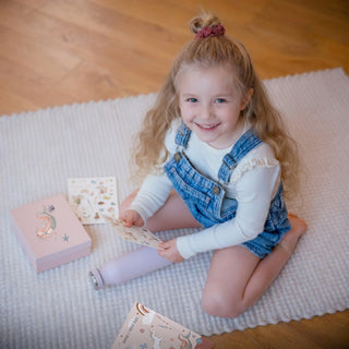 Ein Mädchen sitz auf dem Boden und lächelt glücklich in die Kamera. Sie hält einen Fantasy Stickerbogen aus dem Fantasy Sticker-Set in der Hand. Eine Box ist bereits mit einer Meerjungfrau beklebt.