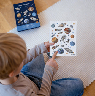 Ein Kind hält ein Stickerbogen in der Hand auf dem die Planeten unseres Sonnensystems, sowie Raketen, ein Astronaut und ein Dackelastronaut inmitten vieler Sterne zu sehen sind.