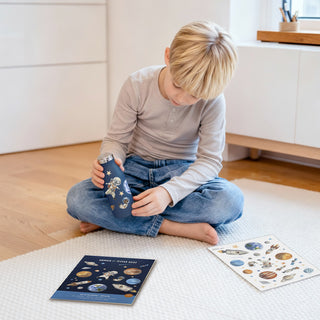 Ein Kind sitzt auf dem Boden und hält eine Trinkflasche, die mit Weltraum-Sticker beklebt ist, in seiner Hand. Es liegt ein Stickerbogen und eine Cardbox von den Kinder Space Sticker auf dem Boden daneben.