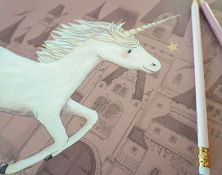 Schreibtischunterlage für Kinder in Dustyrose zeigt ein Einhorn vor einem märchenhaften Schloss, mit zwei pastellfarbenen Stiften.