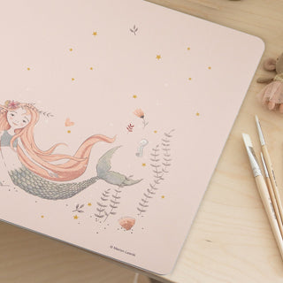 Auf dem Schreibtisch aus Holz liegt eine Kinderschreibtischunterlage mit einer illustrierten Meerjungfrau, Delfin und hübschen Pflanzen.