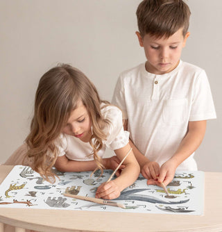Zwei Kinder stehen an einem Kinderschreibtisch und malen mit Pinsel auf einer ABC Schreibtichunterlage mit Tieren. Die Tiere sind in natürlichen Farben auf offwhite Hintergrund