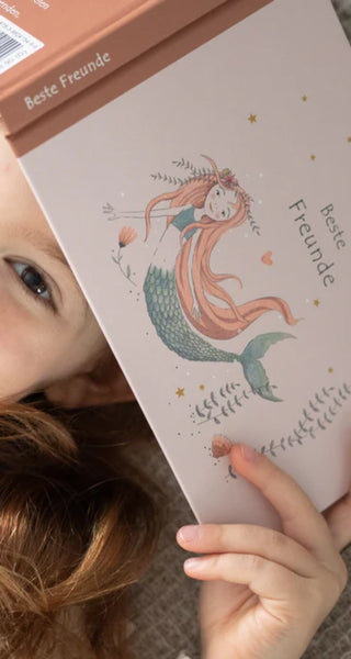 Ein Kind hält ein Buch mit dem Titel "Beste Freunde", darauf eine pastellfarbene Meerjungfrau mit langen Haaren und Blumen.
