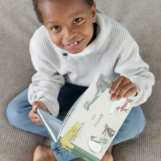 Ein Kind sitzt auf dem Boden, lächelt und hält ein aufgeschlagenes illustriertes Kinderbuch in der Hand, auf dessen Seite verschiedene Tiere abgebildet sind.