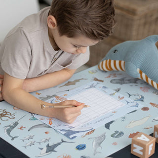 Ein Jungen sitz am Schreibtisch und füllt seinen Stundenplan für die Schule aus.Der Stundenplan liegt auf der Kinderschreibtischunterlage. Diese Unterlage zeigt ganz viele, von Hand illustrierte Meerestiere. Die Grundfarbe ist helles jeansblau. Weiterhilfst ein Stofftier in Form eines Delfin zu sehen.