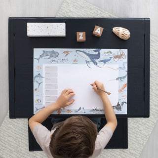 Ein Kind schreibt auf einer Schreibtischunterlage mit Meeresmotiven; Rand mit Meerestieren; Wochenplaner-Layout; Lern- und Geschenkidee für Kinder.