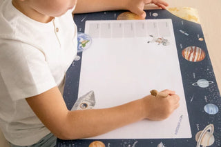 Ein Kind schreibt auf einer Aktivitätsmatte mit Weltraummotiv, illustrierten Planeten und Astronauten, und benutzt einen Stift.